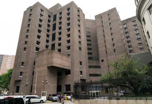 Tall concrete building with numerous windows and adjacent parked cars.